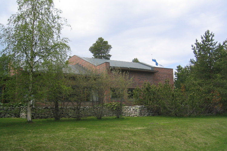 Srk-keskus idästä käsin, matala kivimuuri, edessä nurmea,puita ja pensaita, taustalla liehuu Suomen lippu.